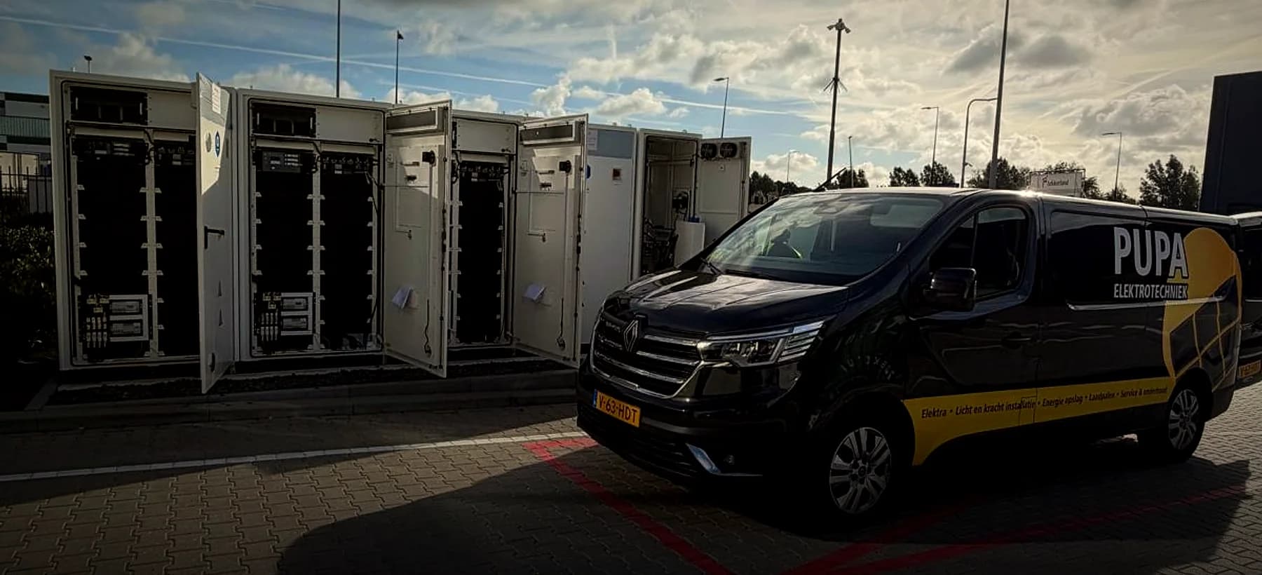 De bus van Pupa Elektrotechniek naast een rij batterijkasten op een bedrijventerrein.
